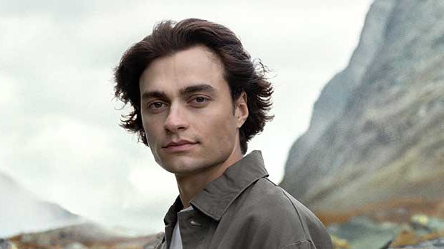 Mann mit schönen Haaren schaut in die Kamera, im Hintergrund wolkenverhangene Berge