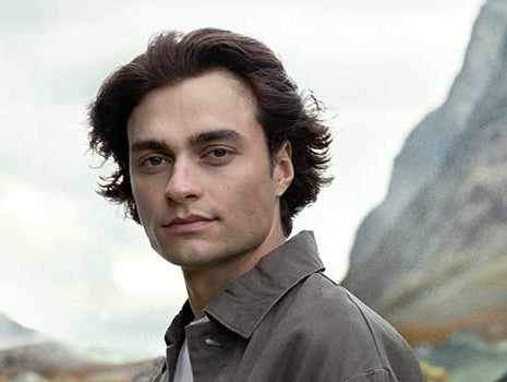 Mann mit schönen Haaren schaut in die Kamera, im Hintergrund wolkenverhangene Berge