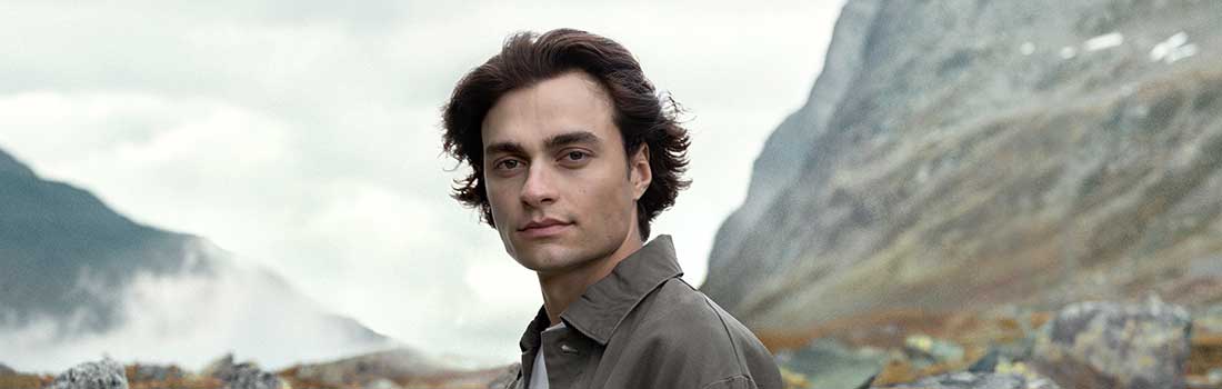 Mann mit schönen Haaren schaut in die Kamera, im Hintergrund wolkenverhangene Berge